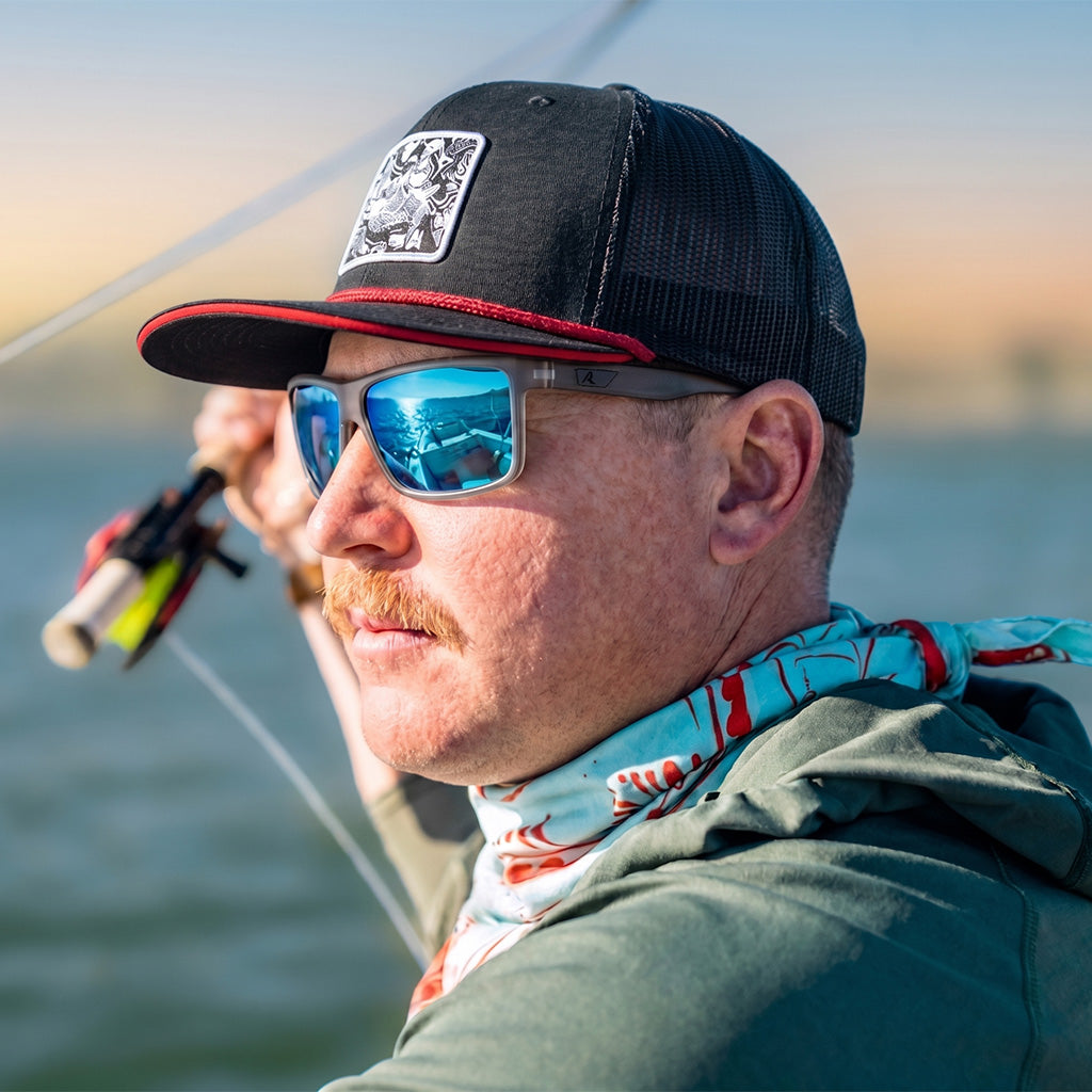 Man wearing a black cap with a logo and sunglasses, holding a fishing rod on a boat.