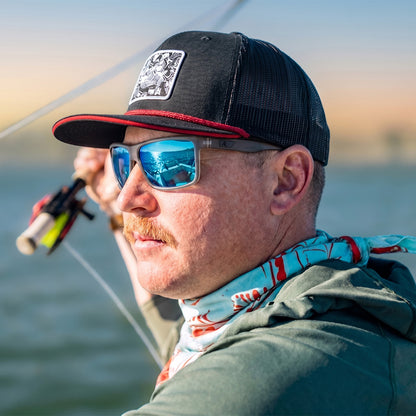 Man wearing a black cap with a logo and sunglasses, holding a fishing rod on a boat.